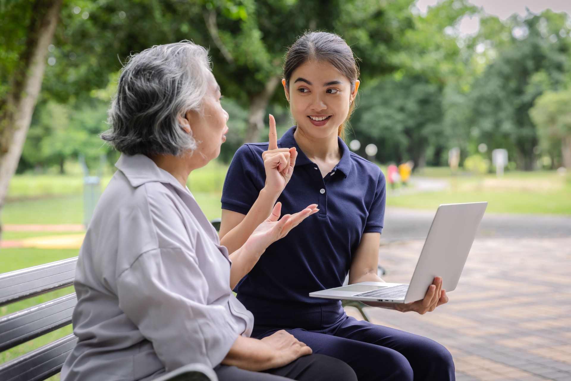 Young caregiver discussing senior care plans with an elderly woman using a laptop while sitting on a park bench, emphasizing attentive support and personalized elderly care in a serene environment.